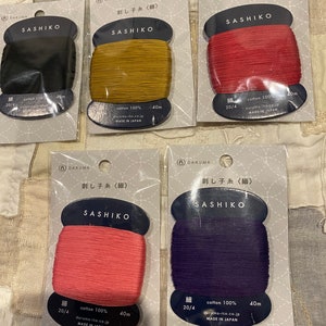May include: Five spools of cotton thread for sashiko embroidery. The spools are labeled "Daruma Sashiko" and are in the colors black, gold, red, pink, and purple. Each spool is 40 meters long and made in Japan.