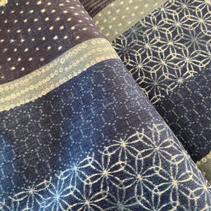 May include: Close-up of folded blue fabric with various patterns. The fabric features geometric designs, including dotted and floral patterns, in shades of blue and white. The texture appears soft and possibly cotton or linen.