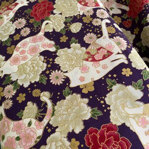 May include: Purple fabric with a repeating pattern of white cats, pink flowers, and gold accents. The cats are depicted with a fan and flowers on their backs. The fabric has a traditional Japanese design.