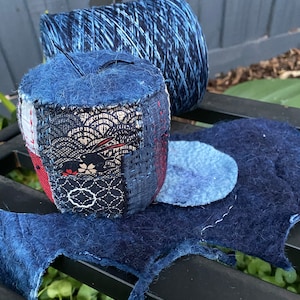 Op de afbeelding: Een handgemaakt blauw speldenkussen met een naald, met patchworkstof met bloemen- en geometrische patronen. Een spoel blauw garen en een viltblauwe mat met een cirkelvormig accent zijn ook zichtbaar, wat wijst op een naai- of knutselthema.