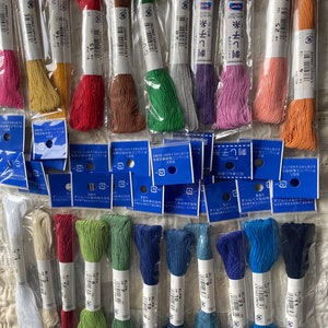 Op de afbeelding: Een assortiment van borduurgaren spoelen in verschillende kleuren, waaronder rood, groen, blauw en bruin. De spoelen zijn verpakt in doorzichtig plastic met blauwe en witte etiketten waarop "オリムパス" en "刺し子糸" staat.