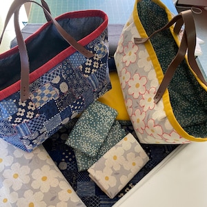 Może przedstawiać: Dwie torby typu tote z skórzanymi uchwytami. Jedna torba ma wzór patchwork w odcieniach niebieskiego, z czerwonym wykończeniem. Druga torba ma kwiatowy wzór w kolorze białym i żółtym na szarym tle. Widoczne są również kawałki materiału.