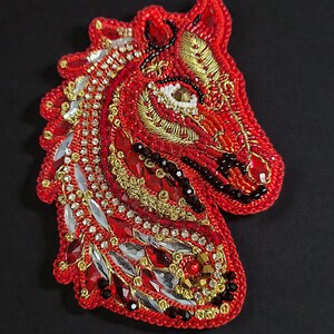 May include: A red, beaded horse head brooch. The brooch features intricate beadwork in shades of red, gold, and clear crystals. The horse's eye is a white bead. The brooch is on a black background. The word "OLCHE" is at the bottom.