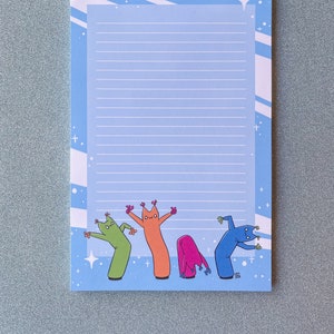 Peut inclure: Bloc-notes bleu à lignes avec une bordure blanche et des étoiles. Le bas du bloc-notes présente une ligne de cinq personnages gonflables en tube de bande dessinée de différentes couleurs, avec le texte "Lora  2023" dans le coin inférieur droit.