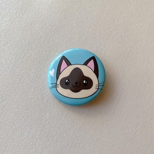 May include: A blue button with a cartoon Siamese cat with brown ears and a brown face on it. The cat has white fur around its eyes and a small white heart above its head.