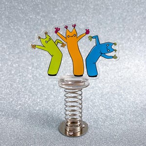 Könnte beinhalten: Ein klarer Acryl-Anhänger mit drei Cartoon-Figuren in Grün, Orange und Blau. Jede Figur hat einen einzigartigen Ausdruck und ist auf einer silbernen Federbasis montiert. Die Figuren haben rosa Hände und ein kleines "owo"-Gesicht.