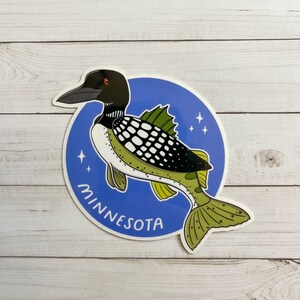 May include: A blue and white sticker with a cartoon loon holding a fish. The loon is black and white with a red eye. The sticker says "Minnesota" in a white cursive font.