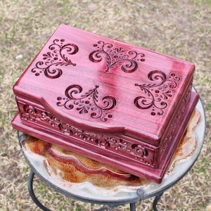 May include: A handcrafted, rectangular wooden box with a deep burgundy finish. The lid and sides feature intricate, carved floral and scrollwork designs. The box is displayed on a small, decorative table.