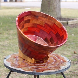 May include: A large wooden bowl with a red, brown, and purple mosaic pattern. The bowl is tilted and sitting on a stand on a table with a red, brown, and white marbled surface.