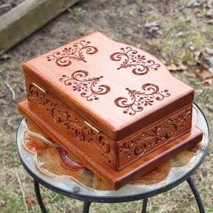 May include: A wooden jewelry box with intricate floral carvings. The box is stained a warm brown color and features decorative carvings on the lid and sides. The box is rectangular with a hinged lid and brass hinges.