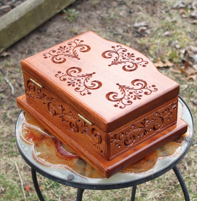 May include: A wooden jewelry box with intricate floral carvings. The box is stained a warm brown color and features decorative carvings on the lid and sides. The box is rectangular with a hinged lid and brass hinges.