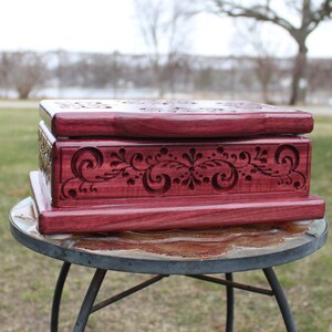 May include: A handcrafted, rectangular wooden box in a deep reddish-purple hue. The box features intricate floral and swirl carvings on the front and top, adding a decorative touch. The box is displayed on a small, round table.