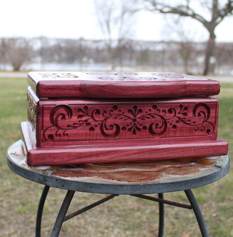 May include: A handcrafted, rectangular wooden box in a deep reddish-purple hue. The box features intricate floral and swirl carvings on the front and top, adding a decorative touch. The box is displayed on a small, round table.