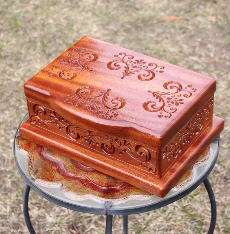 May include: A rectangular wooden box with a rich, reddish-brown finish. The box features intricate, carved floral designs on the lid and sides. It is set on a small, decorative table with a patterned top, showcasing the craftsmanship and detail.