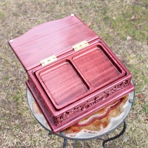 May include: An open, burgundy-colored wooden box with two recessed trays. The box has a hinged lid and ornate carved detailing along the sides. The box is sitting on a glass-topped table.