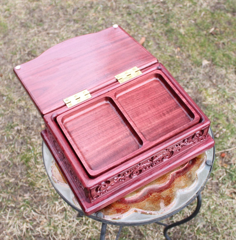 May include: An open, burgundy-colored wooden box with two recessed trays. The box has a hinged lid and ornate carved detailing along the sides. The box is sitting on a glass-topped table.
