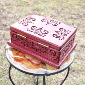 May include: A handcrafted, rectangular wooden box in a deep reddish-purple hue. The box features intricate, carved floral designs on the lid and sides, with brass hinges. The box is displayed on a small, round glass-topped table.