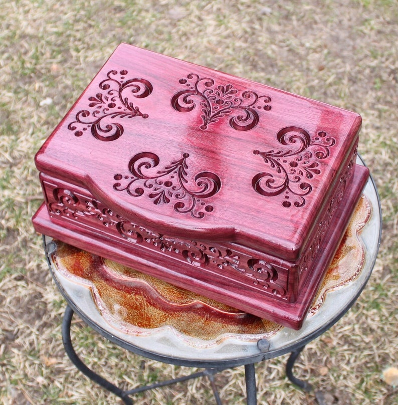 May include: A handcrafted, rectangular wooden box with a deep burgundy finish. The lid and sides feature intricate, carved floral and scrollwork designs. The box is displayed on a small, decorative table.