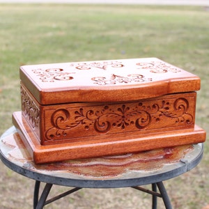 May include: A handcrafted wooden box with intricate floral carvings. The box is stained a warm reddish-brown color and features detailed scrollwork on the lid and sides. The box is rectangular and sits on a matching base, perfect for storing jewelry or keepsakes.
