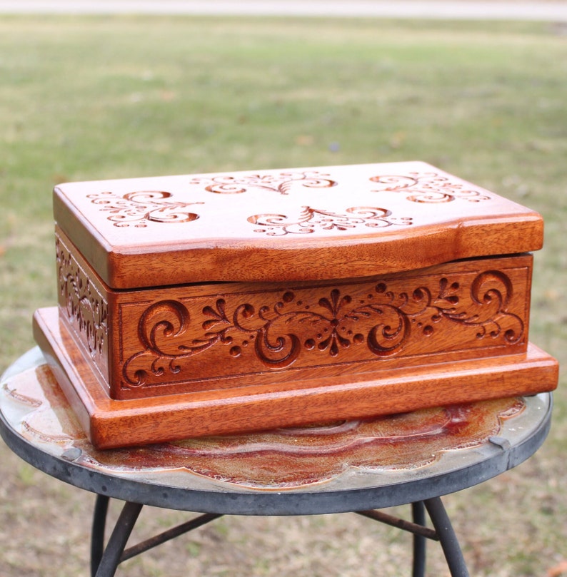 May include: A handcrafted wooden box with intricate floral carvings. The box is stained a warm reddish-brown color and features detailed scrollwork on the lid and sides. The box is rectangular and sits on a matching base, perfect for storing jewelry or keepsakes.