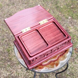 May include: A handcrafted, open wooden box with a deep reddish-purple hue. The box features two recessed trays and ornate carvings along the bottom edge. Brass hinges secure the lid, which is slightly ajar.