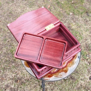 May include: A handcrafted, open wooden box with a deep red hue. The box features a removable tray with two compartments and a hinged lid. The box is made of wood and has a decorative carved design on the sides. The box is sitting on a glass table.