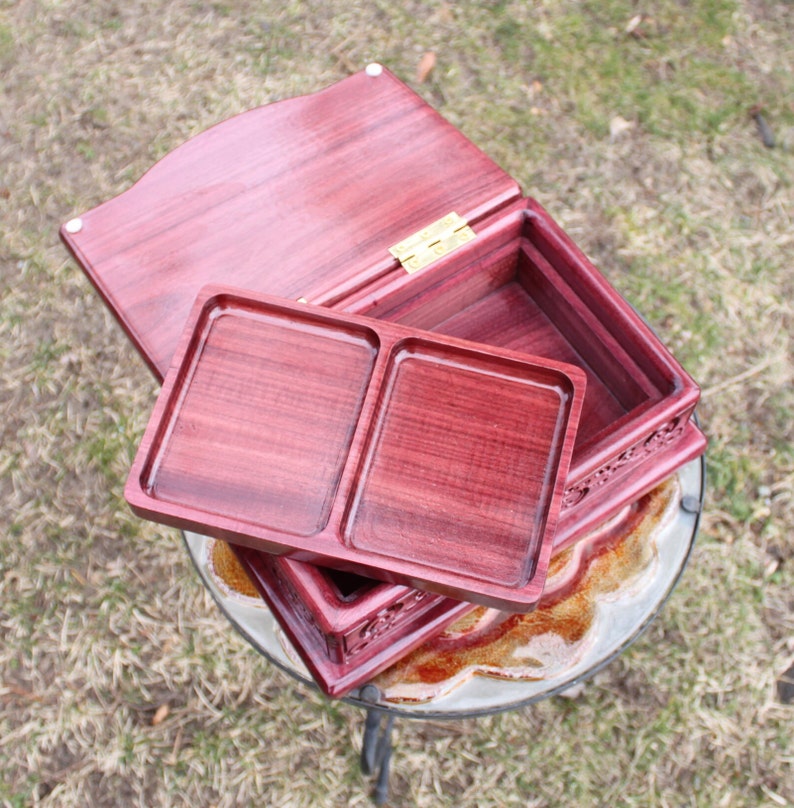 May include: A handcrafted, open wooden box with a deep red hue. The box features a removable tray with two compartments and a hinged lid. The box is made of wood and has a decorative carved design on the sides. The box is sitting on a glass table.