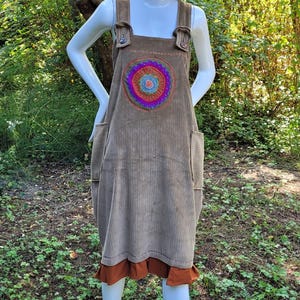 May include: A mannequin wearing a brown corduroy overall dress with a colourful embroidered design. The dress has a rust-coloured ruffle at the hem. A straw hat with a red feather sits atop the mannequin's head. The dress is knee-length.