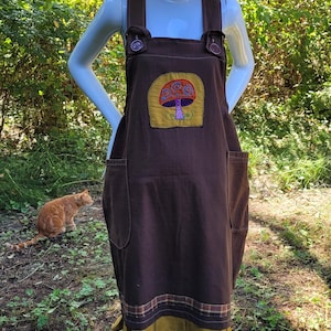 Peut inclure: Robe salopette marron avec un appliqué champignon, portée avec un chapeau de paille. La robe a de grandes poches et des boutons décoratifs. L'ourlet a une garniture plissée de couleur moutarde. Un chat est à l'arrière-plan.