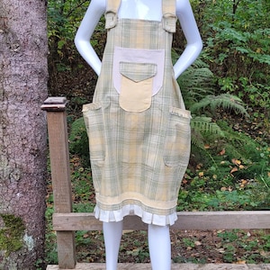 May include: A mannequin wearing a yellow and green plaid overall dress with large pockets and a white frill hem. The mannequin also wears a straw hat with a red flower accent.