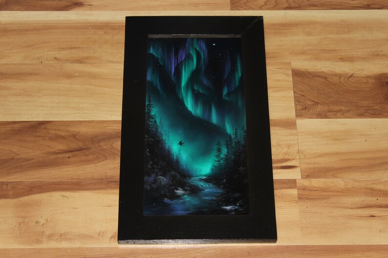 May include: A framed painting of a dark green forest with a river flowing through it. The sky is a deep blue with green and purple aurora borealis lights. A small black bird flies in the sky.