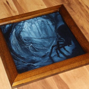 May include: A square wooden frame containing a dark blue and black painting of a forest scene. The artwork depicts a dense, shadowy woodland area with intricate, light-colored spiderwebs stretching across the trees and a large spider silhouette.