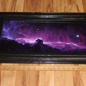 May include: A framed artwork depicting a vibrant nebula scene with shades of purple, pink, and blue, and a black frame. The artwork is approximately 24 inches long. The image includes a wooden ruler.
