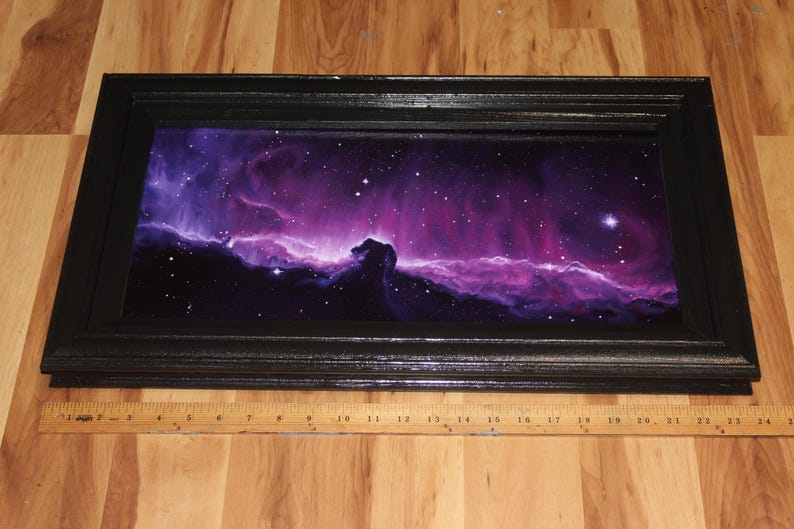 May include: A framed artwork depicting a vibrant nebula scene with shades of purple, pink, and blue, and a black frame. The artwork is approximately 24 inches long. The image includes a wooden ruler.