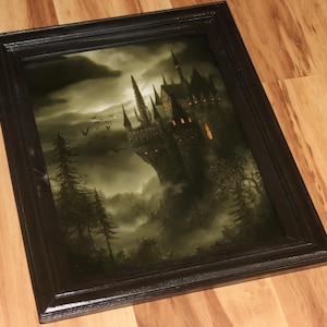 May include: Framed artwork depicting a gothic castle on a misty mountain. The castle is dark with illuminated windows, set against a cloudy sky. The frame is black, and the overall theme is fantasy.