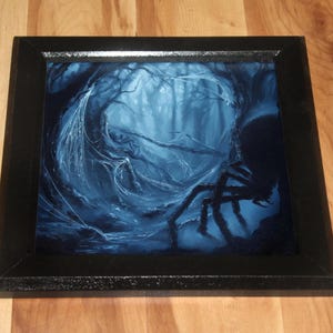 May include: A square framed wall art piece featuring a dark, monochromatic blue illustration of a dense forest scene with intricate spiderwebs draped between trees and a large, dark spider silhouette positioned in the lower right corner.