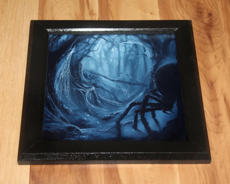 May include: A square framed wall art piece featuring a dark, monochromatic blue illustration of a dense forest scene with intricate spiderwebs draped between trees and a large, dark spider silhouette positioned in the lower right corner.