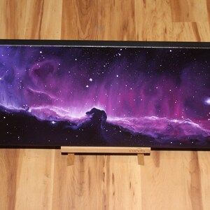 May include: A rectangular canvas print depicting a vibrant nebula in shades of purple and pink, speckled with white stars. The artwork is mounted on a black frame and rests on a wooden stand, showcasing a celestial theme.