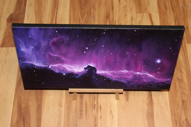 May include: A rectangular canvas print depicting a vibrant nebula in shades of purple and pink, speckled with white stars. The artwork is mounted on a black frame and rests on a wooden stand, showcasing a celestial theme.
