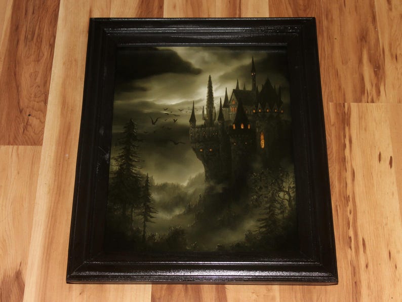 May include: A framed print of a gothic castle on a cliff, set against a dark, stormy sky. The artwork features a detailed depiction of the castle with illuminated windows, surrounded by trees and a misty landscape. The frame is black.
