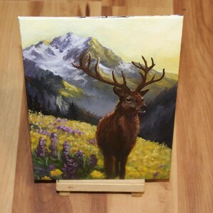May include: An oil painting on canvas shows a stag with large antlers in a field of yellow and purple wildflowers. The background features a mountain range with snow-capped peaks under a yellow sky. The artwork is displayed on a small wooden easel.
