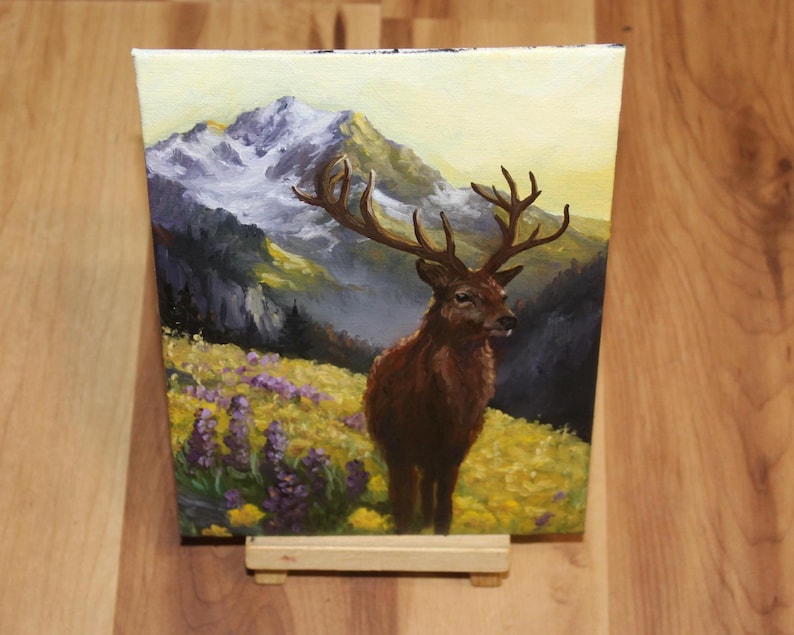 May include: An oil painting on canvas shows a stag with large antlers in a field of yellow and purple wildflowers. The background features a mountain range with snow-capped peaks under a yellow sky. The artwork is displayed on a small wooden easel.