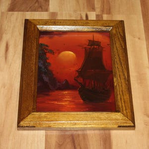 Pu&ograve; includere: Un dipinto incorniciato raffigurante un veliero scuro su un mare rosso vibrante sotto una grande luna luminosa. La scena include una costa rocciosa con fogliame verde scuro sul lato sinistro. L'opera &egrave; racchiusa in una cornice di legno.