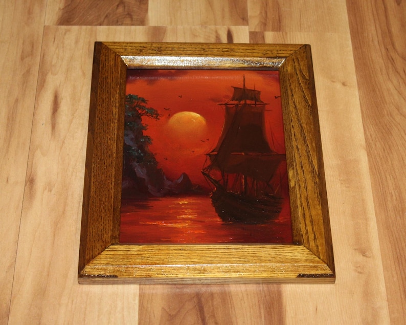Pu&ograve; includere: Un dipinto incorniciato raffigurante un veliero scuro su un mare rosso vibrante sotto una grande luna luminosa. La scena include una costa rocciosa con fogliame verde scuro sul lato sinistro. L'opera &egrave; racchiusa in una cornice di legno.