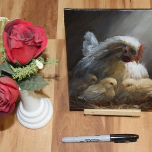 May include: An oil painting on a small easel depicts a hen with three chicks. The hen is white and gray, with a red comb. Two red roses and a white vase are to the left. A black Sharpie marker is at the bottom.