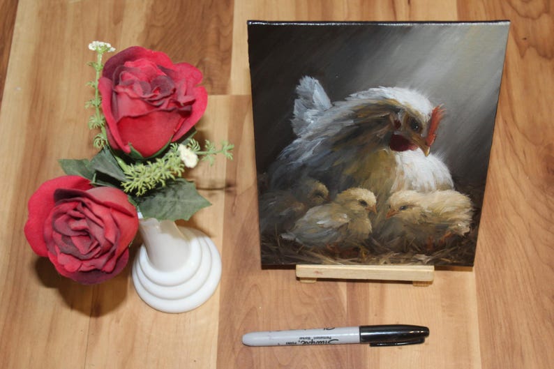 May include: An oil painting on a small easel depicts a hen with three chicks. The hen is white and gray, with a red comb. Two red roses and a white vase are to the left. A black Sharpie marker is at the bottom.