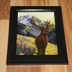 May include: A framed painting depicting a deer with large antlers in a field of yellow and purple flowers, with a mountain range backdrop and a yellow sky. The frame is black.