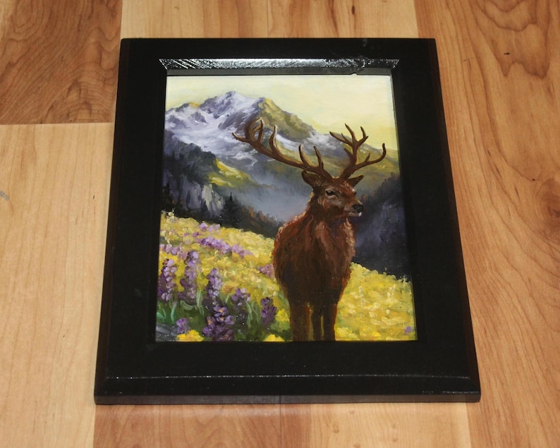 May include: A framed painting depicting a deer with large antlers in a field of yellow and purple flowers, with a mountain range backdrop and a yellow sky. The frame is black.