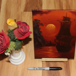 Pu&ograve; includere: Un dipinto rettangolare su tela raffigurante un veliero scuro sull'acqua al tramonto con un grande sole arancione, posizionato accanto a un vaso bianco contenente rose artificiali rosse e gialle e un pennarello Sharpie nero.