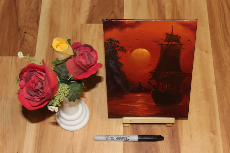 Pu&ograve; includere: Un dipinto rettangolare su tela raffigurante un veliero scuro sull'acqua al tramonto con un grande sole arancione, posizionato accanto a un vaso bianco contenente rose artificiali rosse e gialle e un pennarello Sharpie nero.